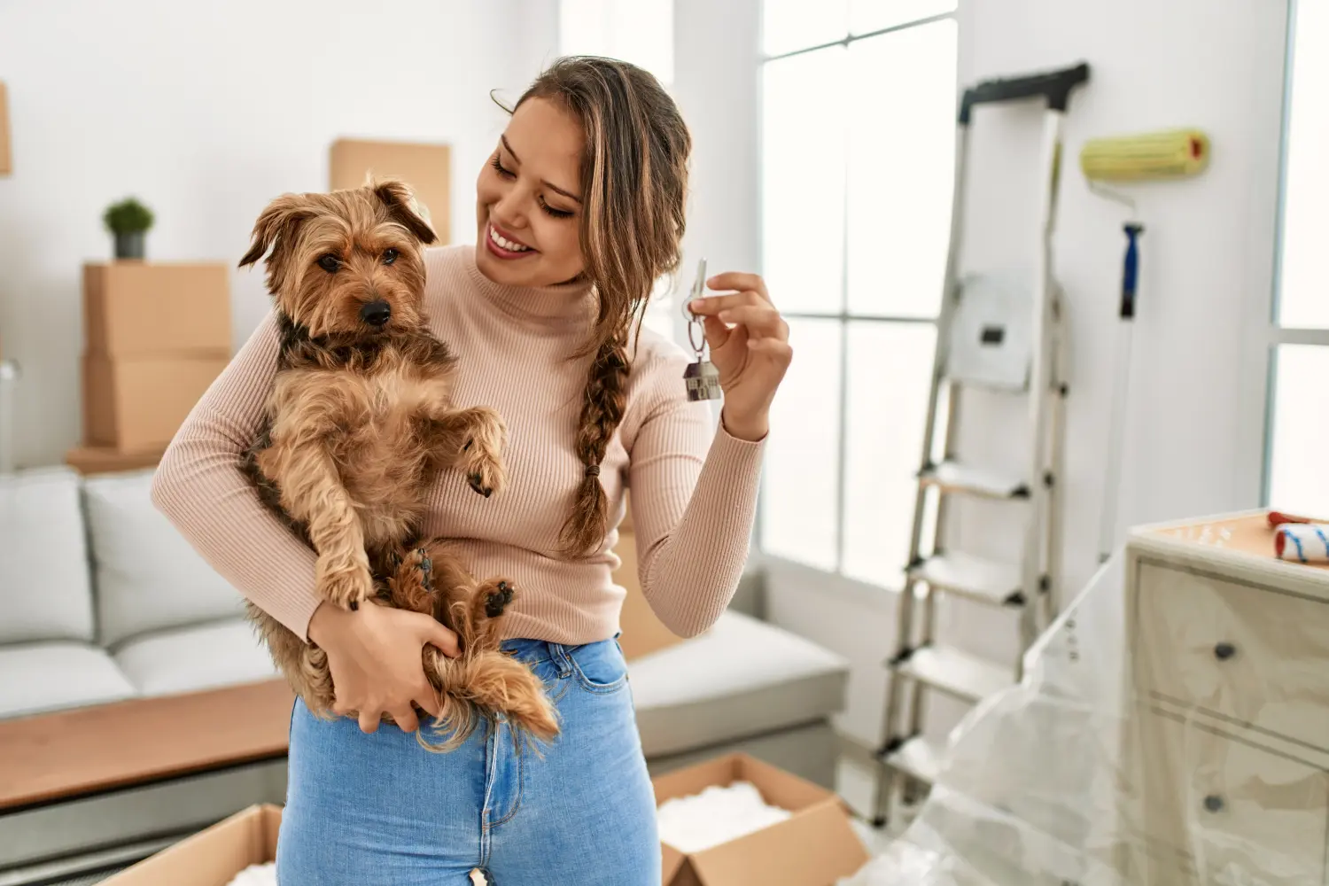 Junge schöne hispanische Frau, die ihren Hund umarmt und den Schlüssel für ihr neues Zuhause hält